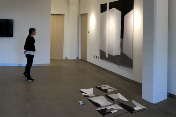 Person stands in a gallery viewing a large abstract, geometric painting of a white cube with gray and black background.