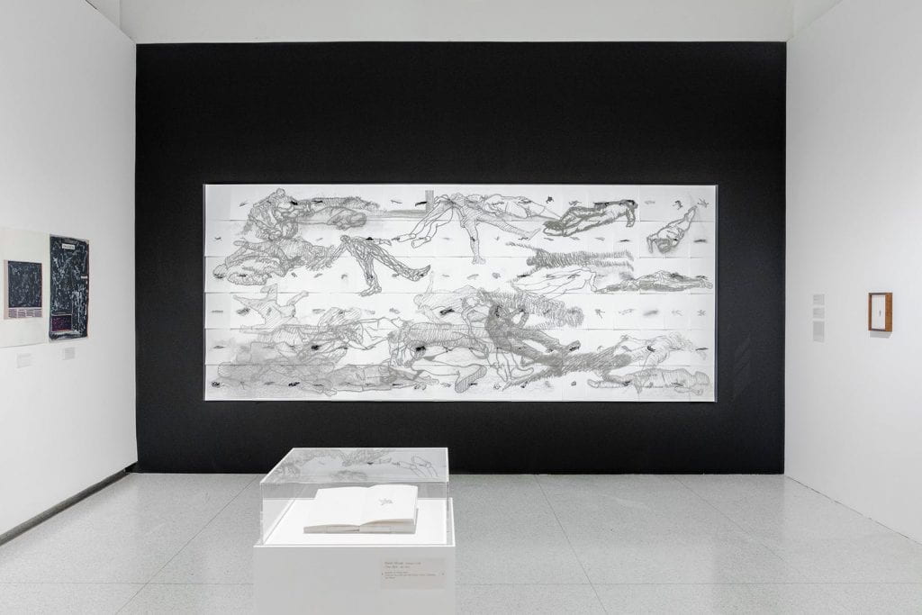 Gallery view of large drawing on a black wall with a book on a pedestal in the foreground.