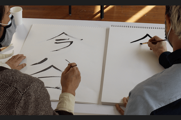 Two people paint on a sketchbook with black ink.